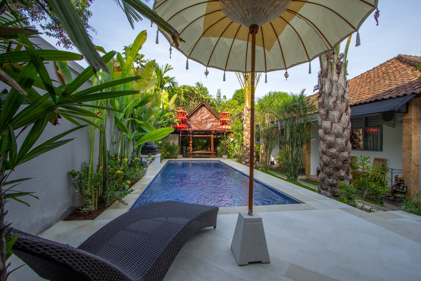 A luxurious swimming pool area with traditional Balinese architecture, featuring a decorative umbrella for shade, lounger seating, lush tropical vegetation, and ornate temple structures in the background. The modern white deck contrasts beautifully with the blue water and natural surroundings.