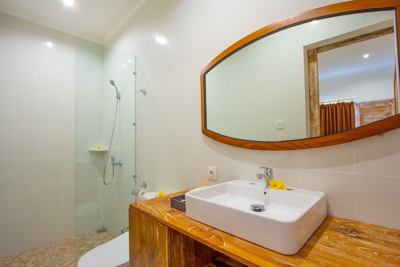 Modern bathroom with wooden vanity, white vessel sink, large arched mirror with wooden frame, glass shower enclosure, white tile walls, and yellow flower decorations
