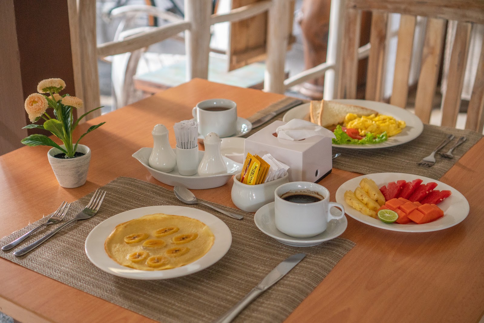 Elegantly arranged breakfast table with pancakes, fresh fruit platter, corn, coffee, and morning beverages. Decorated with fresh flowers and white tableware on a wooden table with neutral placemats.
