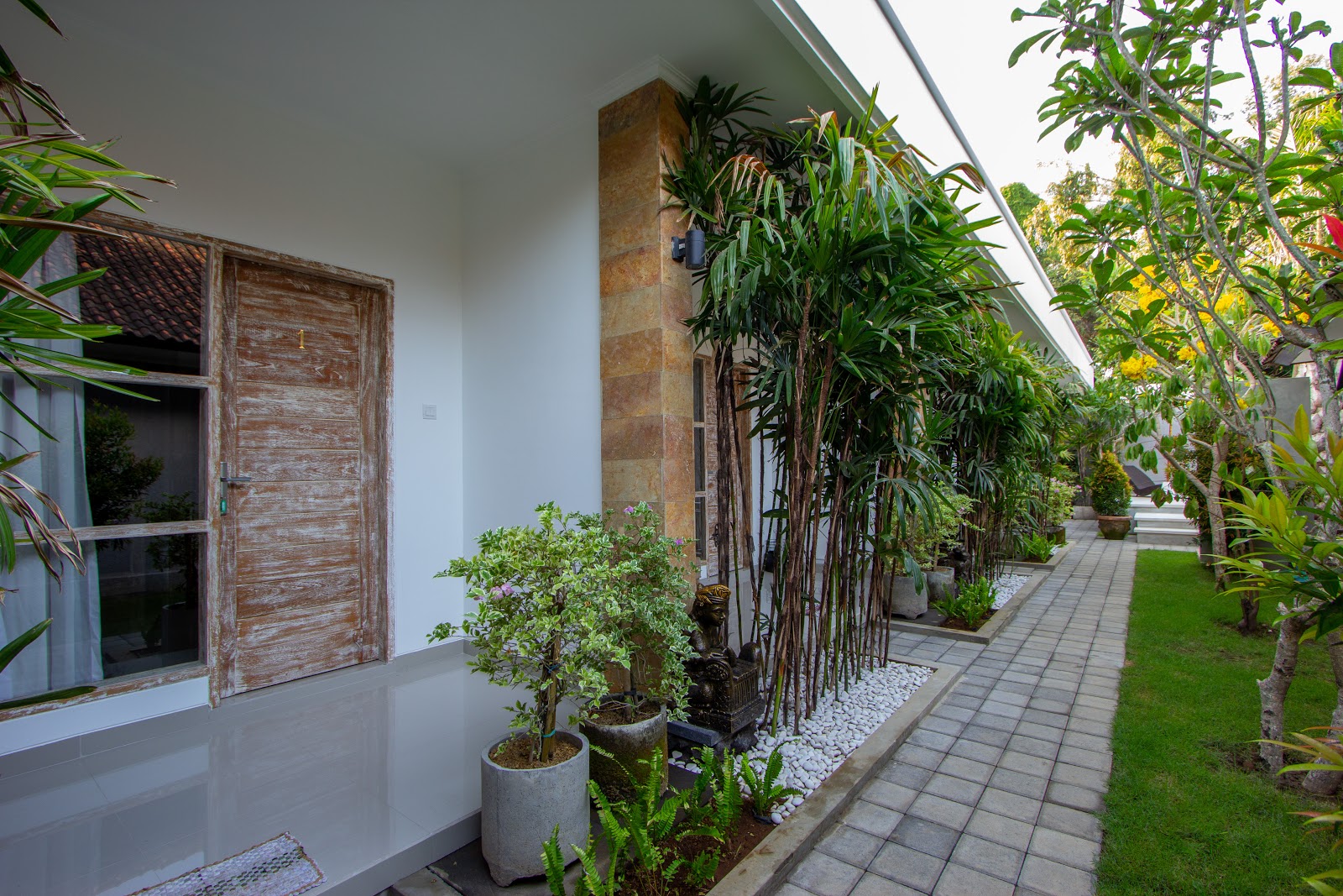 A well-maintained exterior walkway featuring white walls, wooden doors, lush tropical plants, and manicured lawn. The corridor shows modern minimalist design with natural greenery, creating an inviting and serene atmosphere typical of upscale Balinese accommodations.